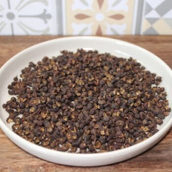 Grains de poivre de Sichuan dans une assiette blanche sur une table en bois, fond de motifs colorés.