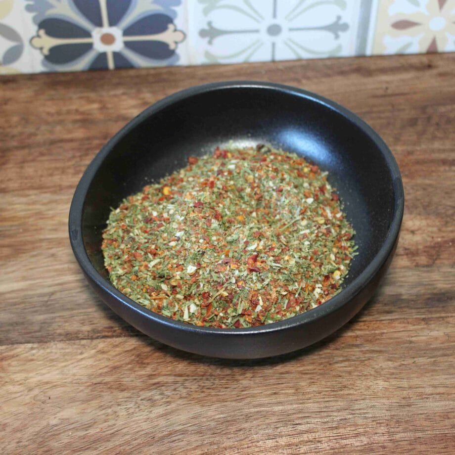 Assortiment coloré d'herbes et d'épices dans un bol noir sur une table en bois, fond de carreaux décoratifs.