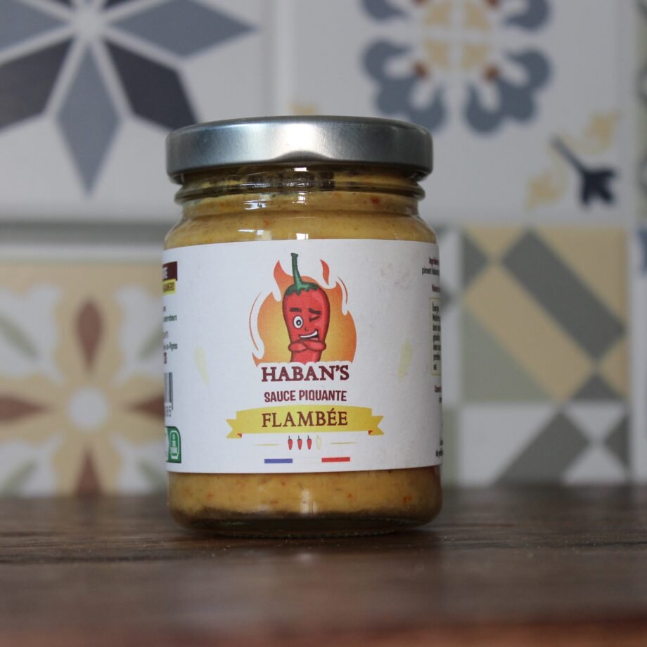 Bocal de sauce piquante Haban's flambée posé sur une table en bois avec motif de fond géométrique.