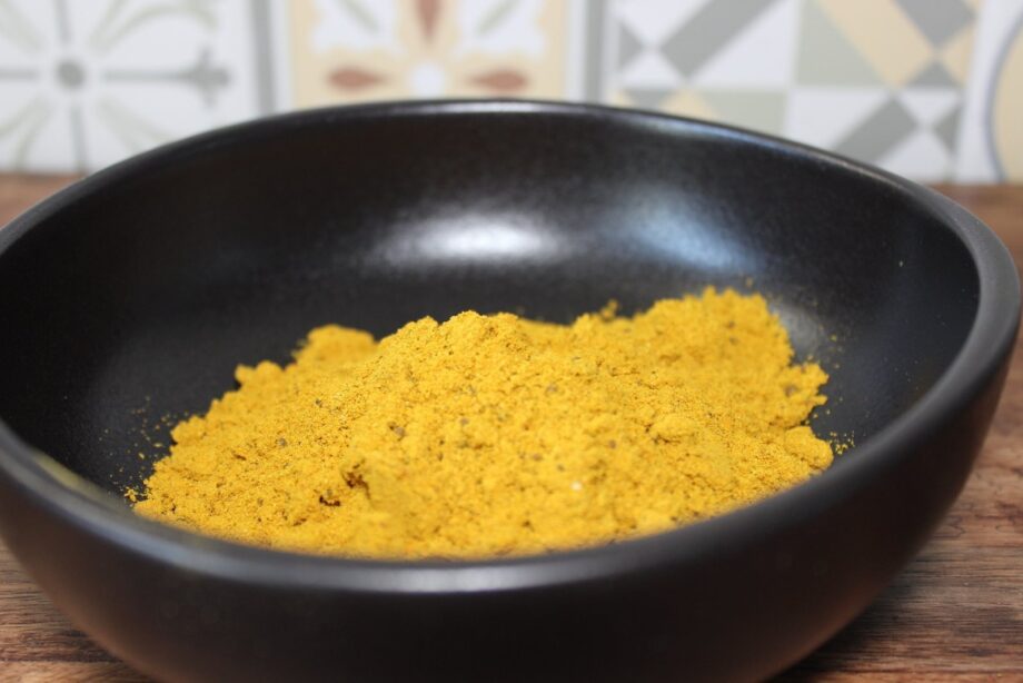 Poudre de curry jaune dans un bol noir sur table en bois, avec carrelage en arrière-plan.