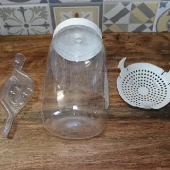Kit de fermentation maison : bocal en plastique, couvercle spécial et panier filtre sur un plan de travail en bois.