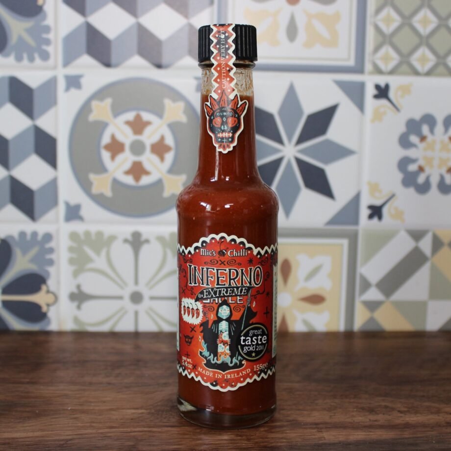 Bouteille de sauce piquante Inferno Extreme Chili posée sur une table en bois devant un mur en carreaux colorés.