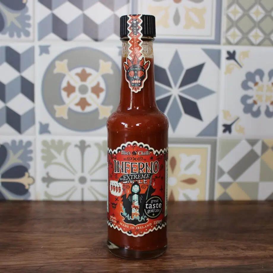 Bouteille de sauce piquante Inferno Extreme Chili posée sur une table en bois devant un mur en carreaux colorés.