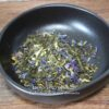 Mélange de thé vert et fleurs séchées dans un bol noir sur table en bois, prêt pour une infusion parfumée et colorée.