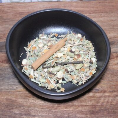 Bol d'épices diverses sur une table en bois, incluant une branche de cannelle, cardamome et herbes séchées.