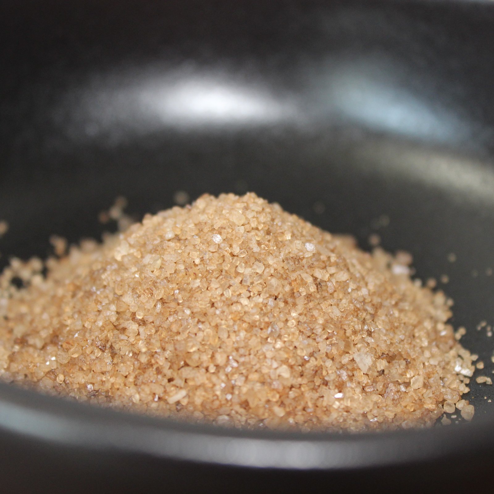 Tas de sucre brun cristallisé dans un bol noir, éclairé délicatement pour en montrer la texture granuleuse.