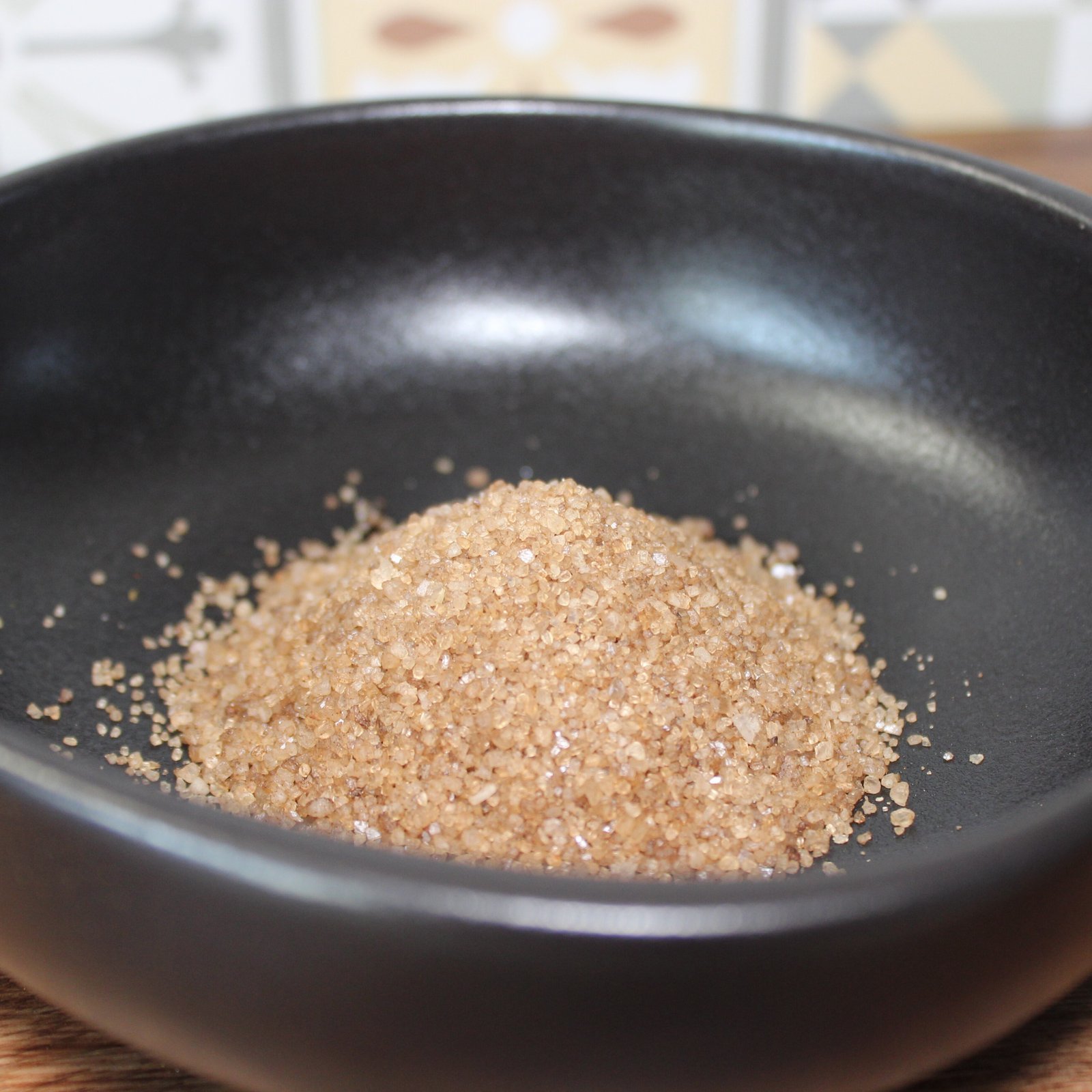 Sucre de canne brun dans un bol noir, idéal pour pâtisserie et boissons. Texture granuleuse et couleur riche.