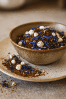 Bol de thé en vrac aux fleurs bleues et ingrédients naturels, sur assiette en céramique.