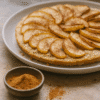 Tarte aux pommes maison avec des tranches saupoudrées de cannelle, accompagnée d'un bol de cannelle en poudre.