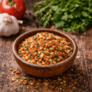 Mélange d'épices colorées dans un bol en terre cuite sur table en bois avec tomates, ail et herbes en arrière-plan.