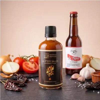 Bouteille de sauce piquante et bière artisanale entourées de légumes frais sur ardoise noire.